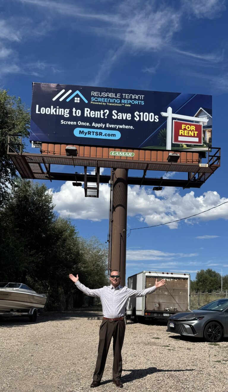 Man in a checkered shirt stands in a gravel lot with arms raised beneath a large billboard advertising rental services.